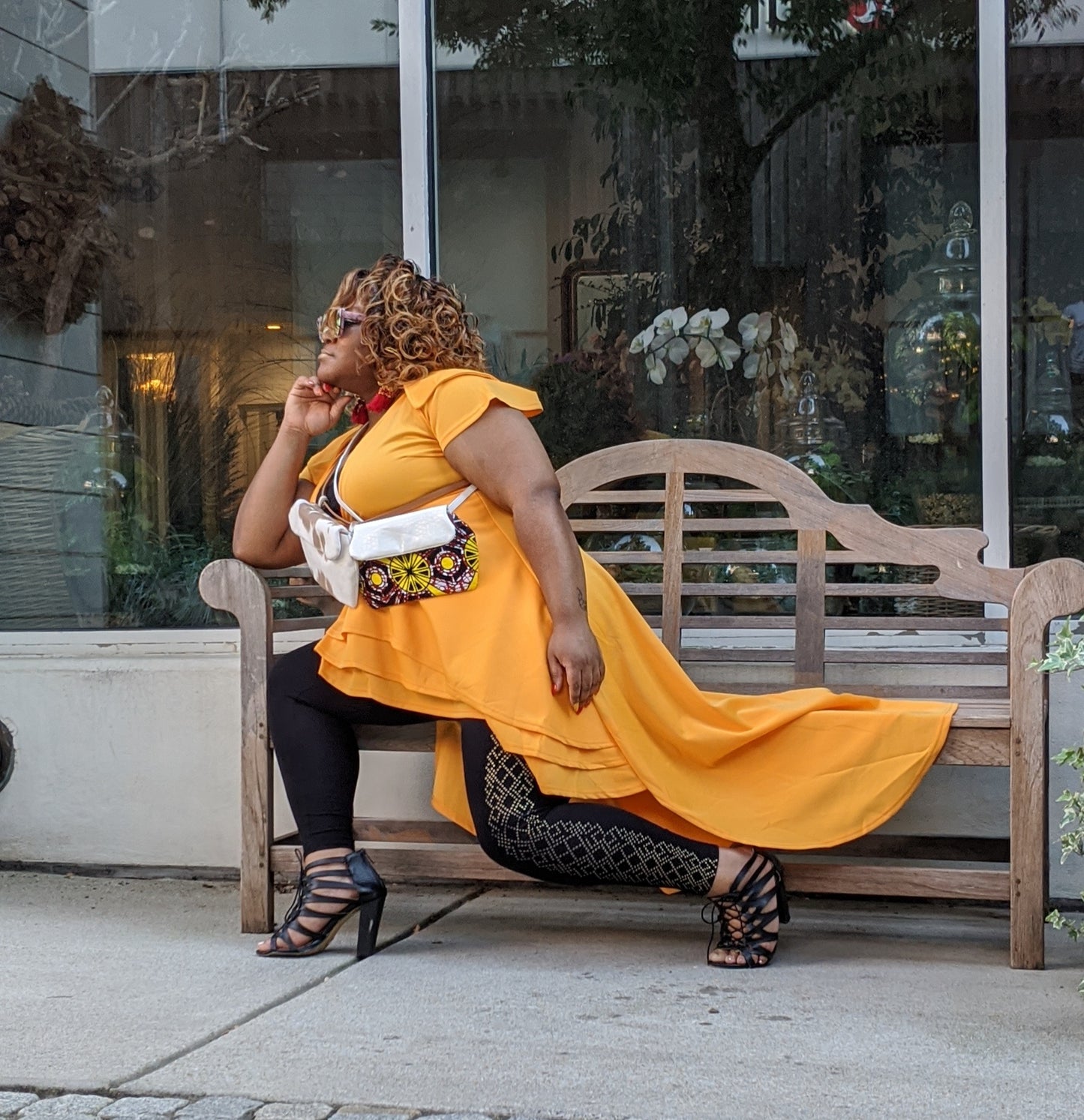 Curvy black woman sitting sideways on bench outside wearing flowing goldenrod dress, two crossbody bags and heels